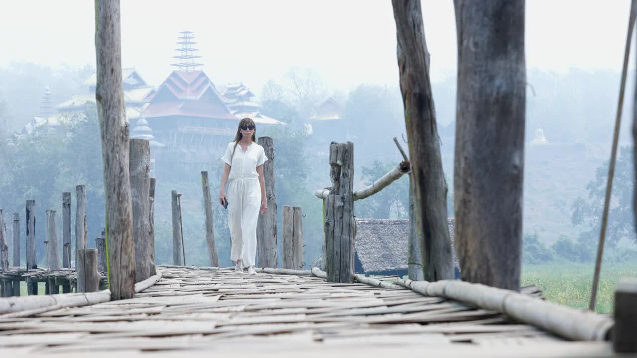 在雾蒙蒙的早晨，一名身穿白衣的年轻女子走在一座木桥上，逐渐向镜头靠近视频下载