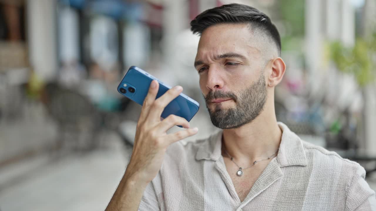 年轻的西班牙裔男子坐在咖啡店露台的桌子上用智能手机发送语音信息视频素材