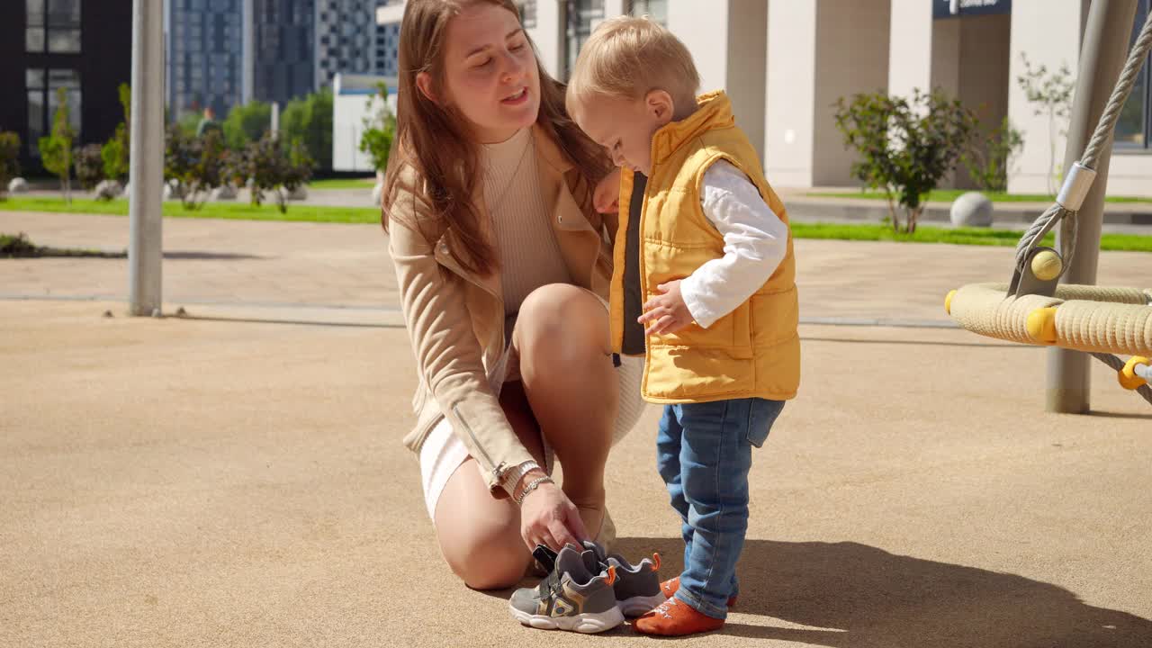 年轻的妈妈在操场上帮她的小儿子穿鞋。孩子们在户外玩耍，孩子们在户外，暑假和假期视频素材