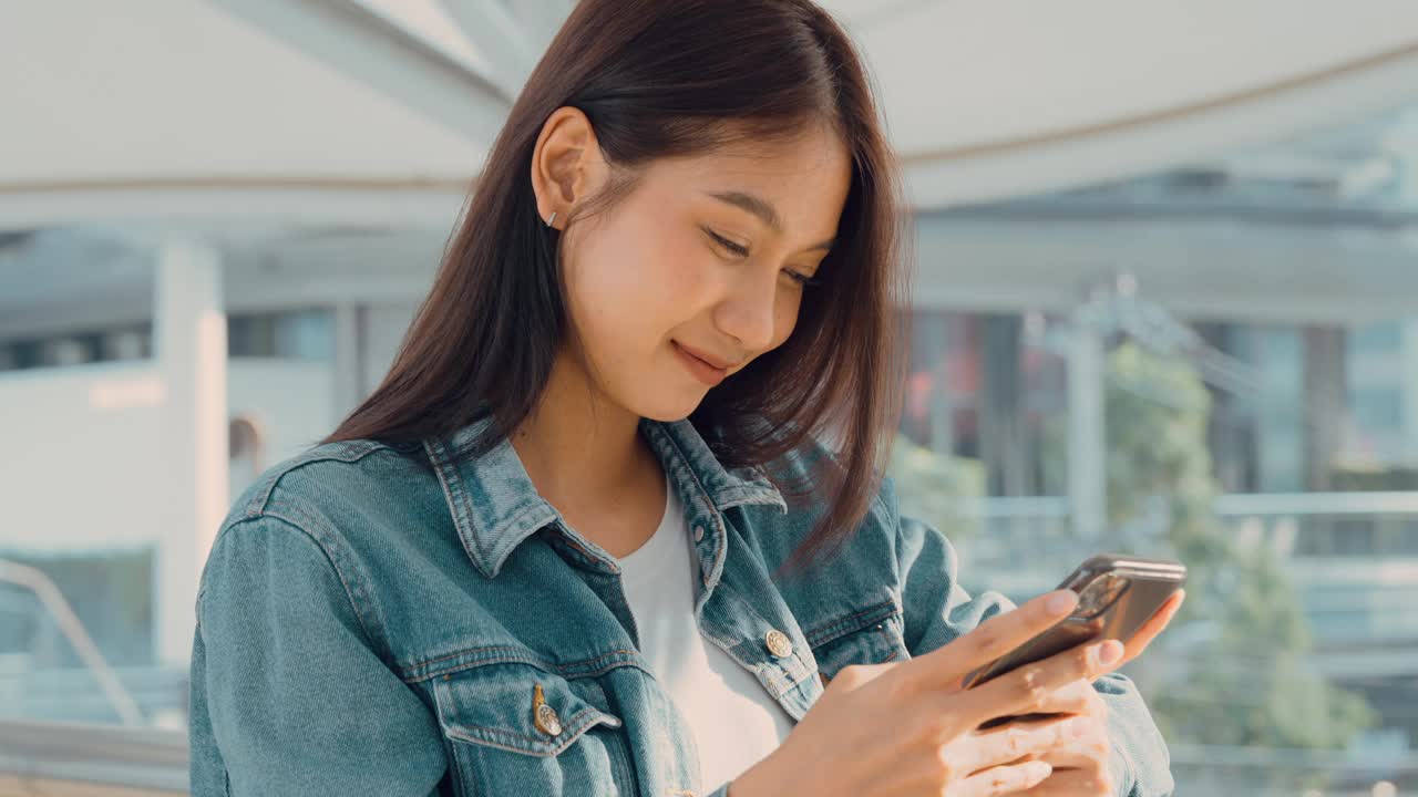 年轻的亚洲女子站在城市里，在街上使用手机视频素材