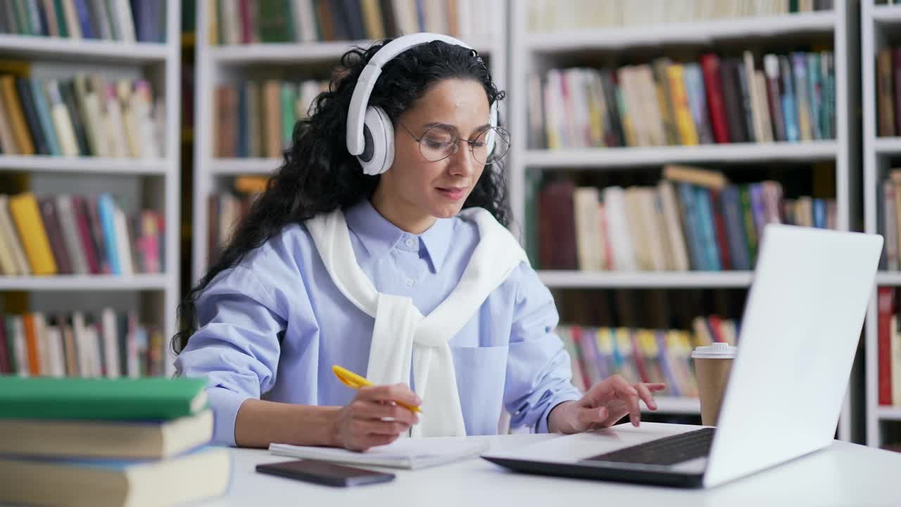 女学生戴着耳机在校园图书馆听在线电子学习视频课程。视频素材