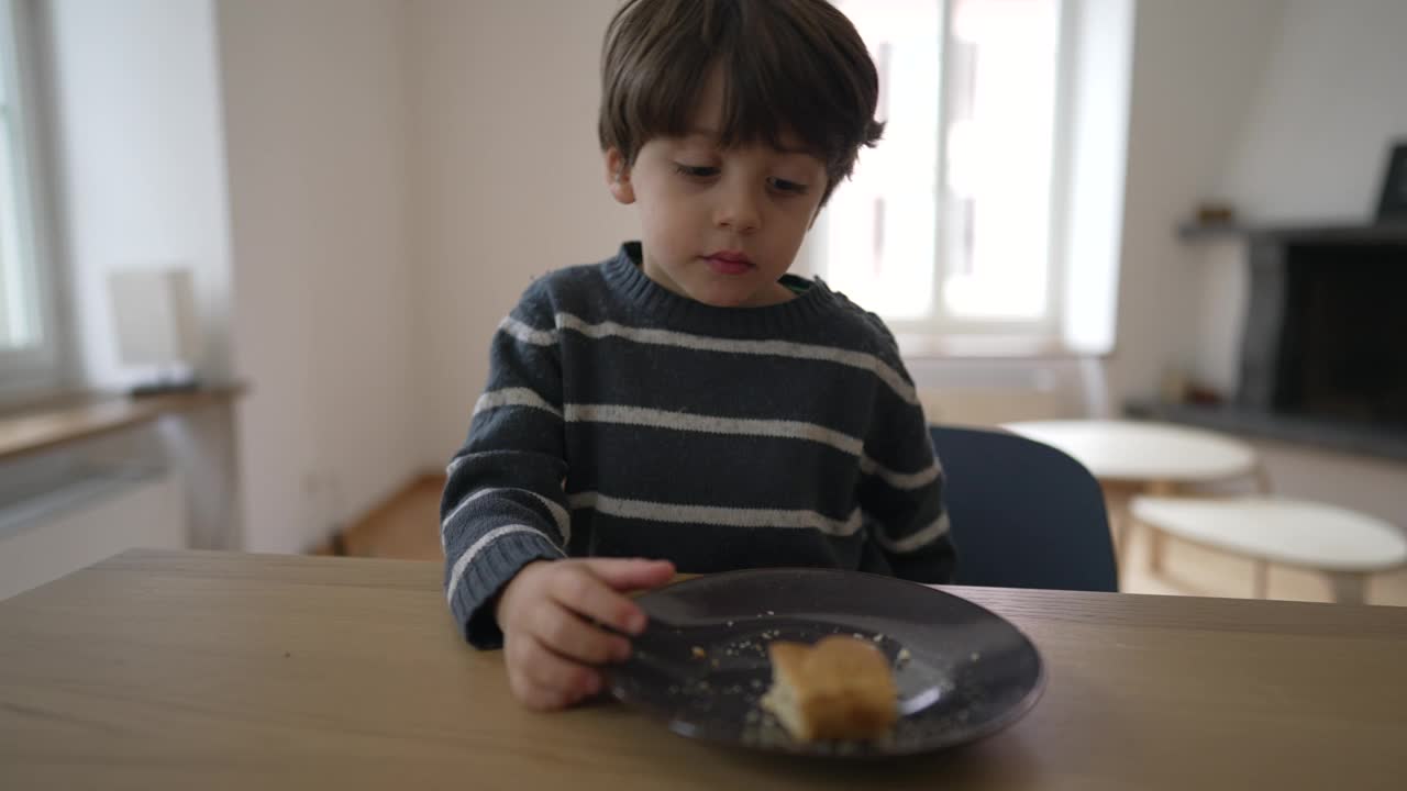 一个小男孩在吃面包，一个四岁的小男孩坐在室内的桌子旁吃着碳水化合物食物视频素材