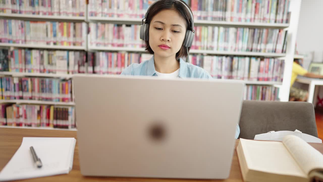 亚洲女大学生在图书馆戴着耳机，在笔记本电脑上上网学习，在图书馆为考试而学习视频素材