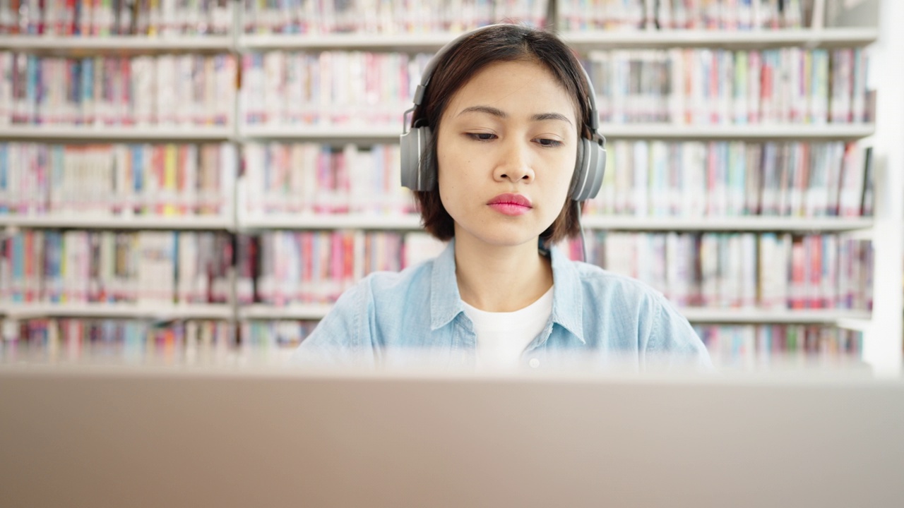 亚洲女大学生在图书馆戴着耳机，在笔记本电脑上上网学习，在图书馆为考试而学习视频素材