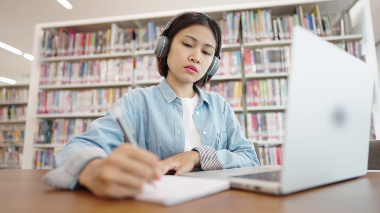 亚洲女大学生在图书馆戴着耳机，在笔记本电脑上上网学习，在图书馆为考试而学习视频素材