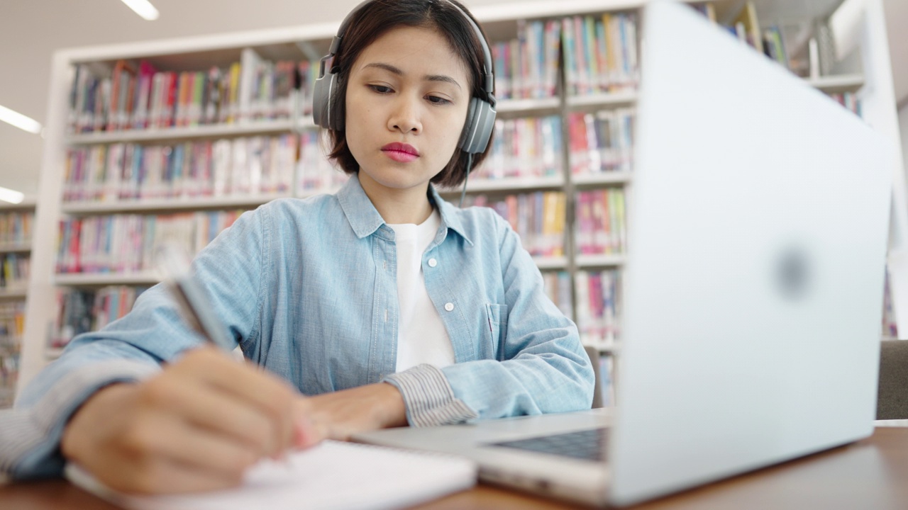 亚洲女大学生在图书馆戴着耳机，在笔记本电脑上上网学习，在图书馆为考试而学习视频素材