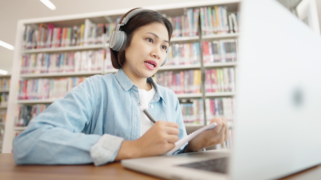 亚洲女大学生在图书馆戴着耳机，在笔记本电脑上上网学习，在图书馆为考试而学习视频素材