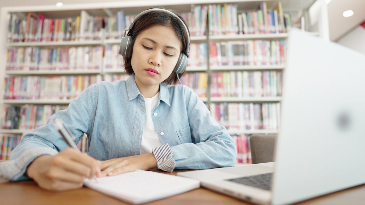 亚洲女大学生在图书馆戴着耳机，在笔记本电脑上上网学习，在图书馆为考试而学习视频素材