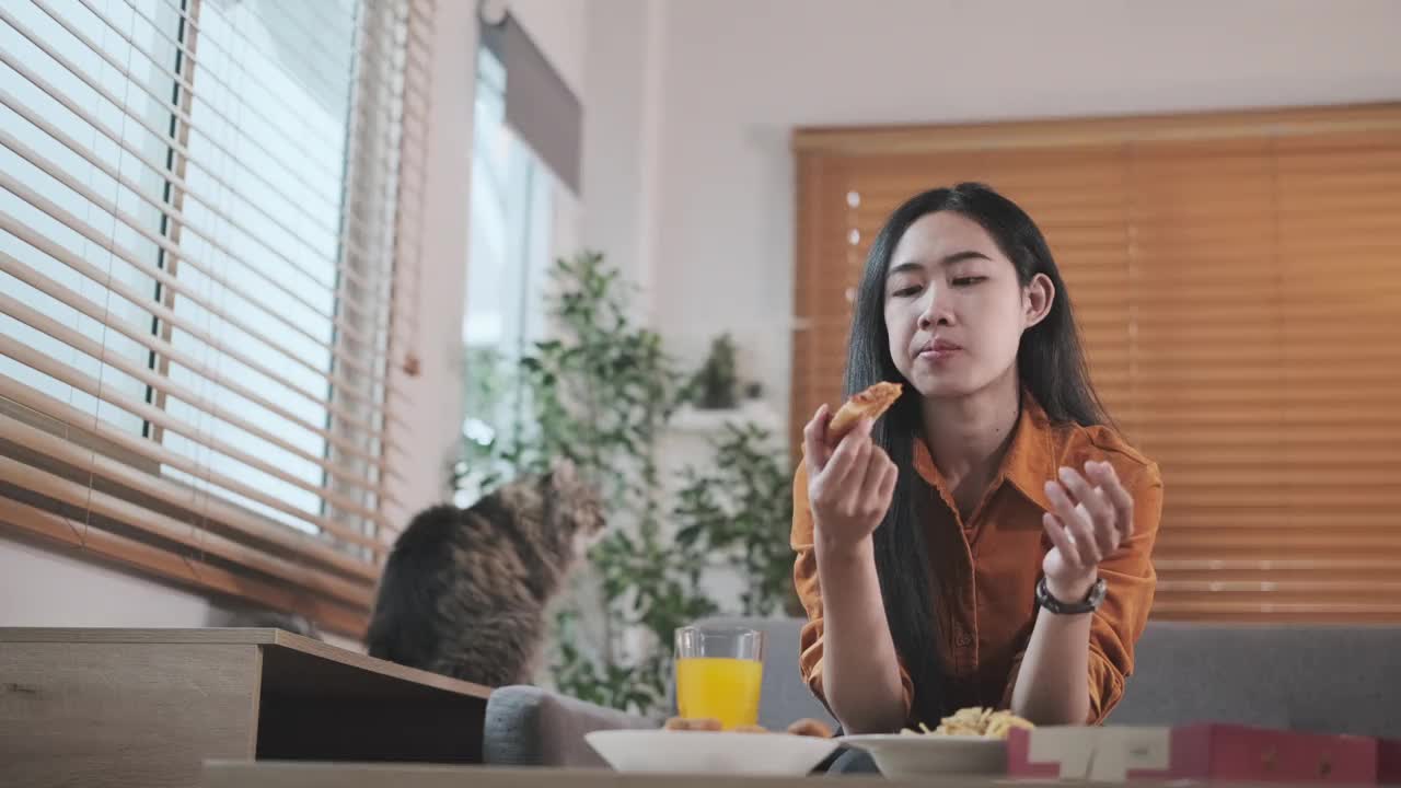 美丽的亚洲女孩拿着一片披萨，咬着它，坐在舒适的客厅的沙发上。视频素材