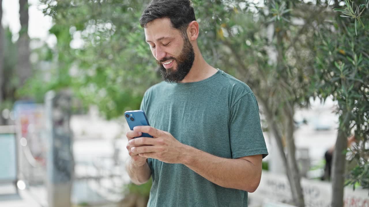 年轻的西班牙裔男子在公园用智能手机微笑视频素材