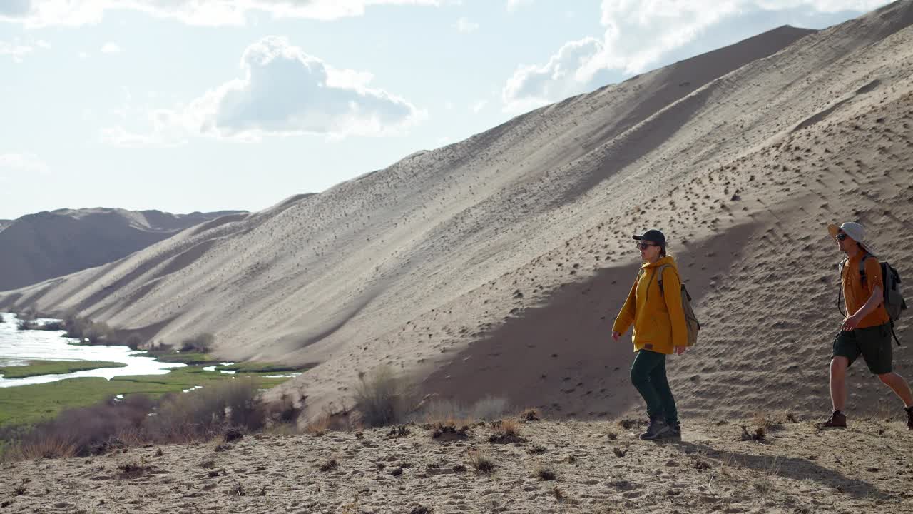 两个年轻的徒步旅行者沿着沙漠地区的侧视图走到目的地视频素材