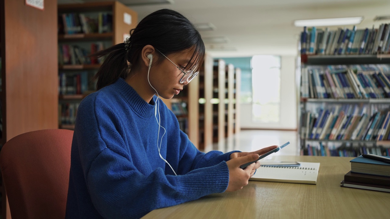 女学生戴着眼镜和耳机，在图书馆里用平板电脑敲击屏幕。视频素材