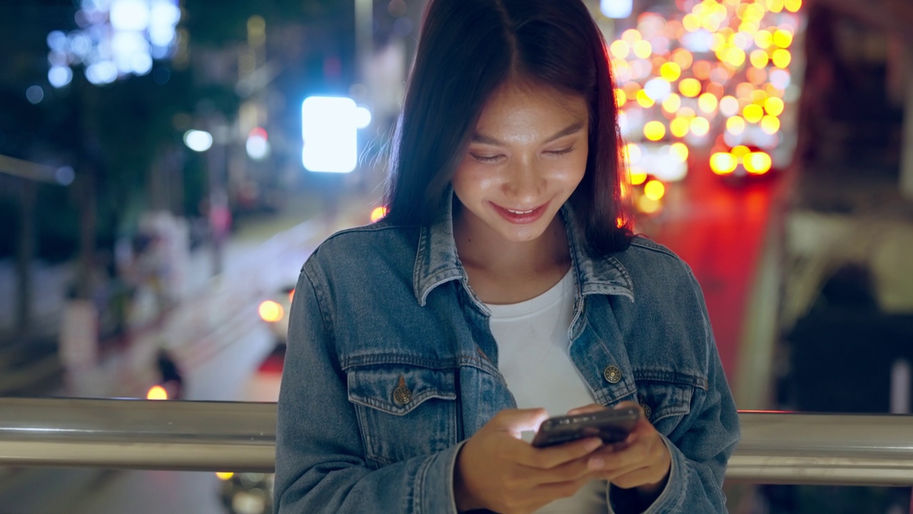 美丽的年轻亚洲女子站在城市街道在晚上使用手机。一个快乐的女人站在那里用手机打字视频素材