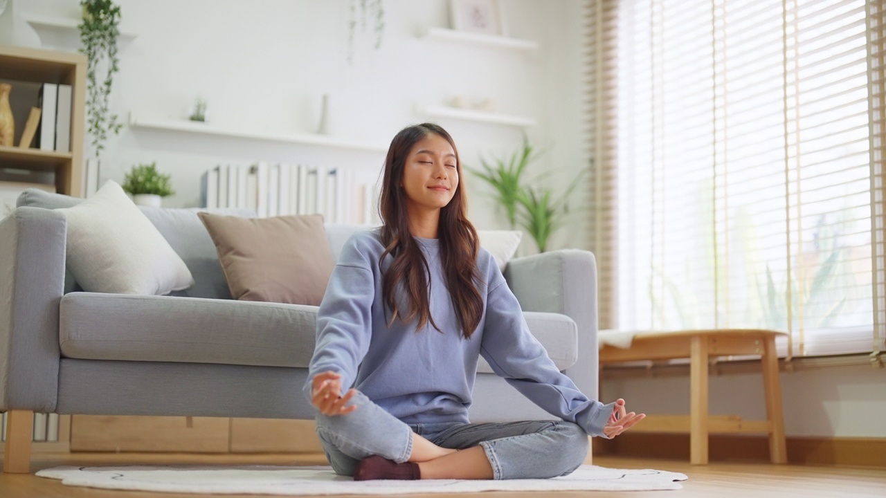 快乐的年轻亚洲女性在家里练习瑜伽和冥想，坐在客厅的地板上，以莲花的姿势，闭上眼睛放松。正念冥想和幸福概念视频素材