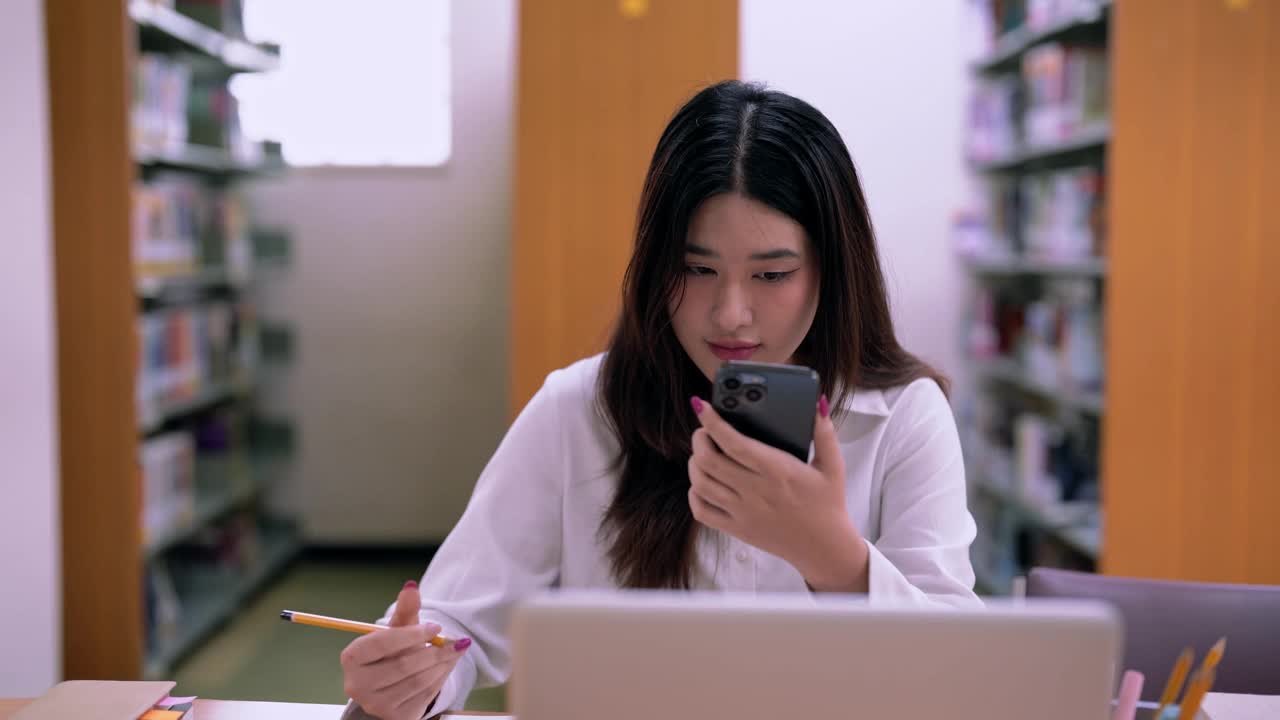 亚洲女学生在图书馆用智能手机做笔记，成人教育理念。视频素材