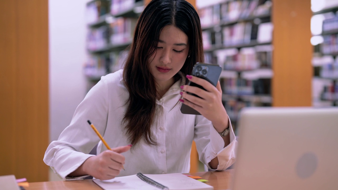 亚洲女学生在图书馆用智能手机做笔记，成人教育理念。视频素材