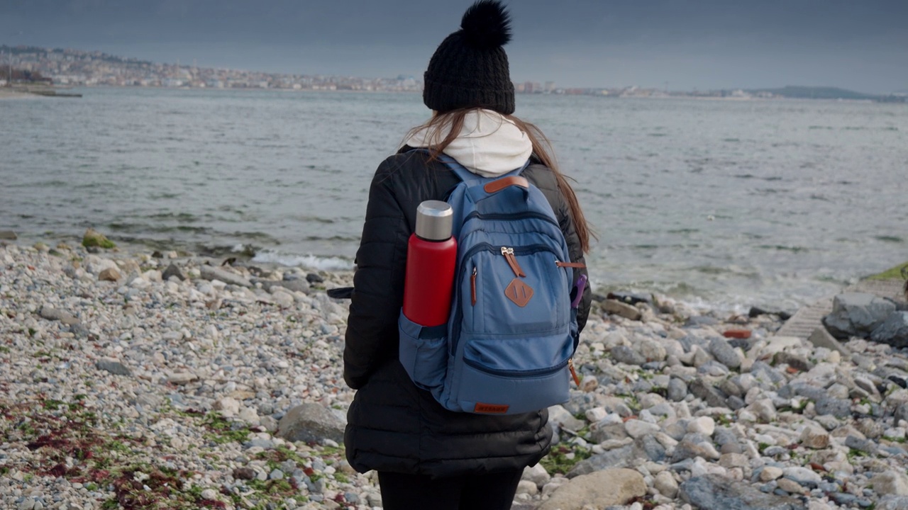 年轻的女徒步旅行者背着背包和保温瓶在寒冷多云和刮风的日子里走在岩石海滩上。徒步探险、探险、旅游、旅行的概念视频素材