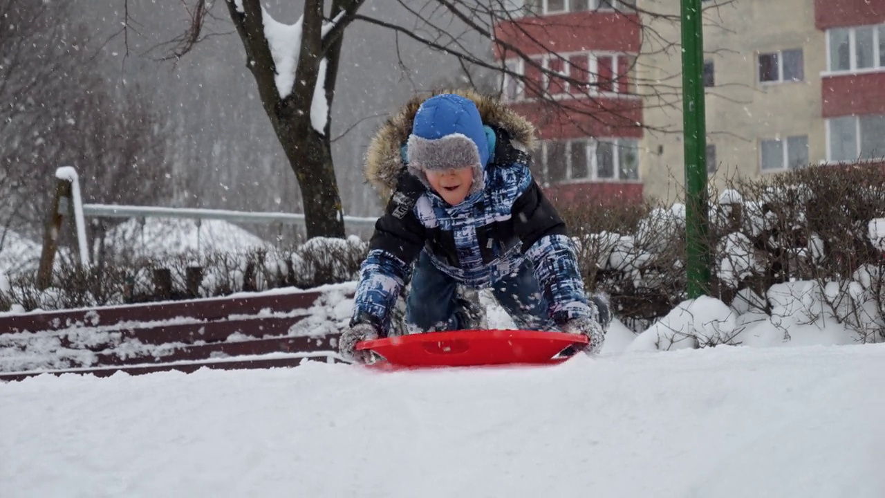 一个小男孩带着灿烂的笑容，坐在塑料雪橇上滑下雪坡，镜头以慢动作拍摄，增强了运动和动作的感觉视频素材