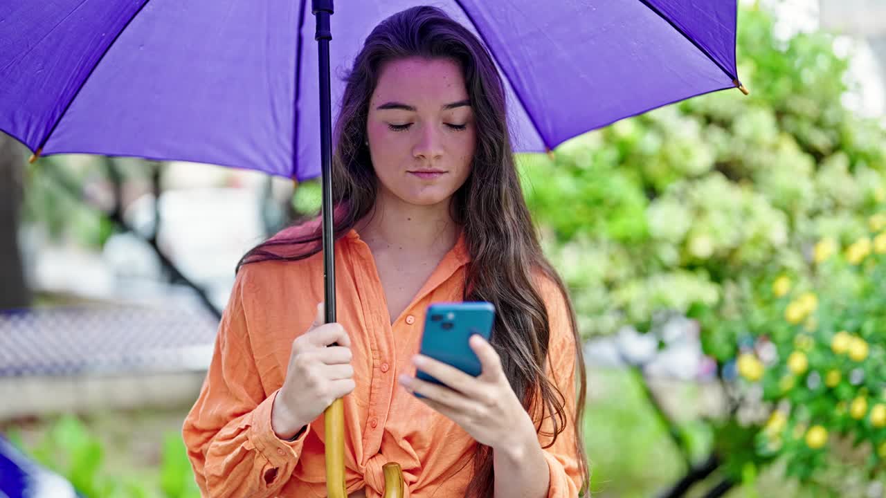 年轻美丽的西班牙女子在公园用智能手机撑伞视频素材