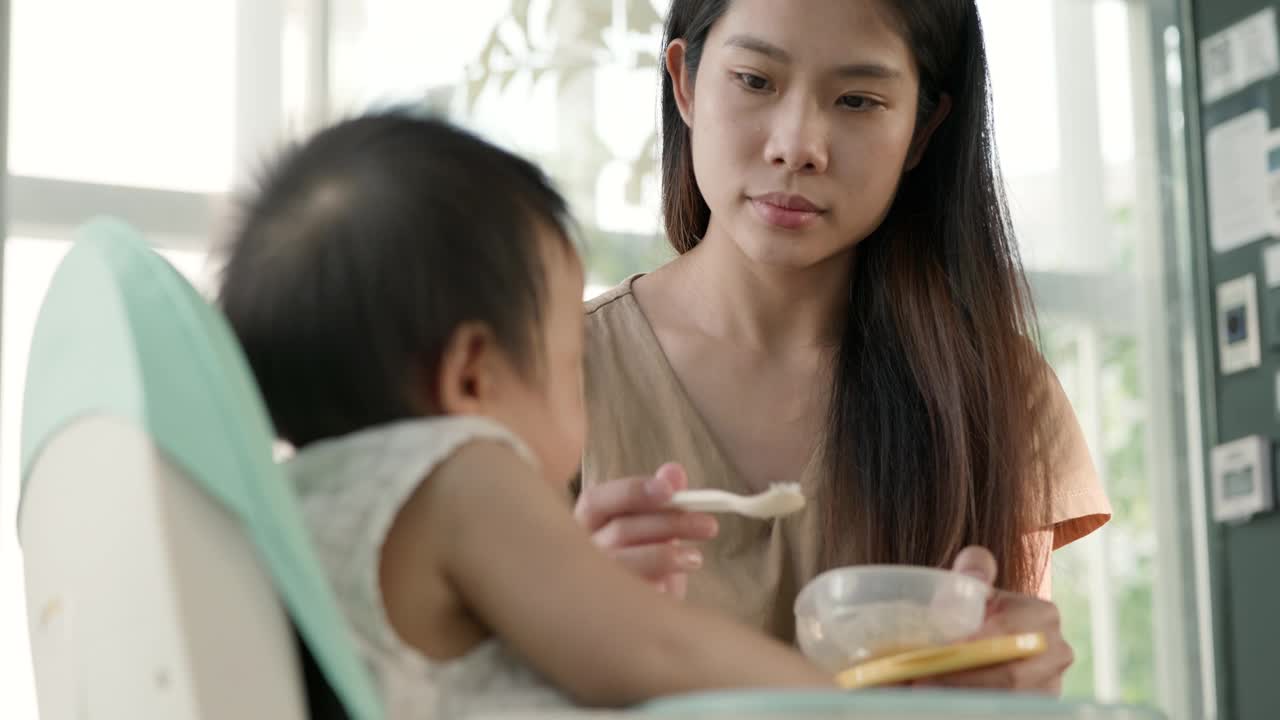 亚洲母亲坐在家里的高脚椅上给女婴喂早餐。视频素材