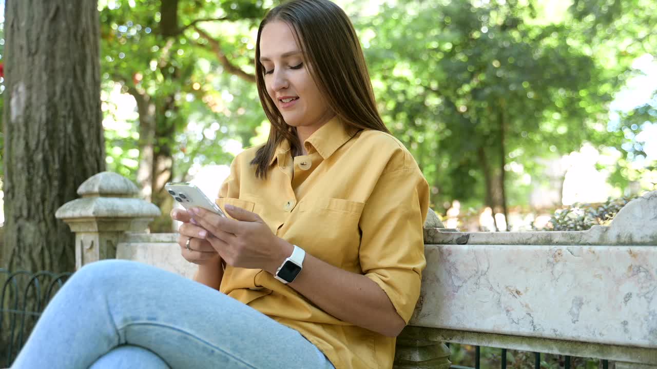 漂亮开朗的女人坐在公园里用智能手机，微笑的女学生拿着手机，滚动新闻，在网上发短信视频素材