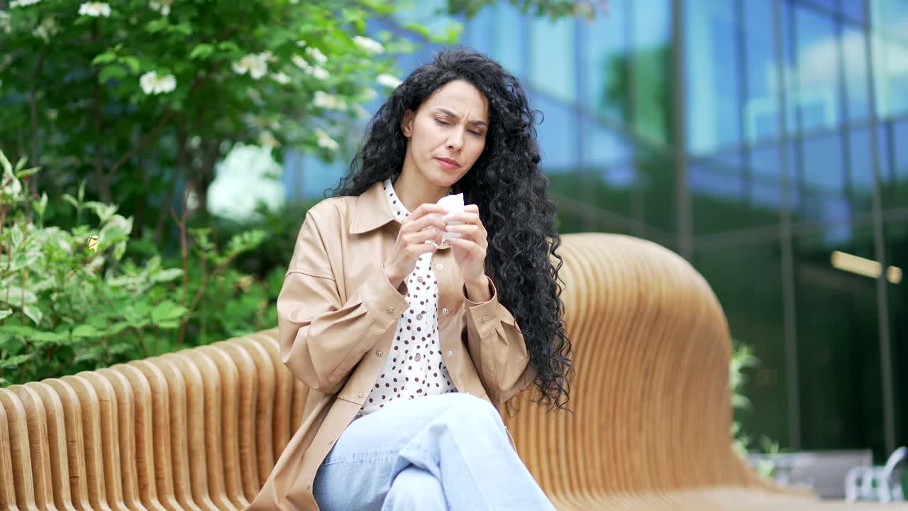 生病的年轻女员工坐在办公大楼附近街道的长凳上，患有季节性过敏症视频素材