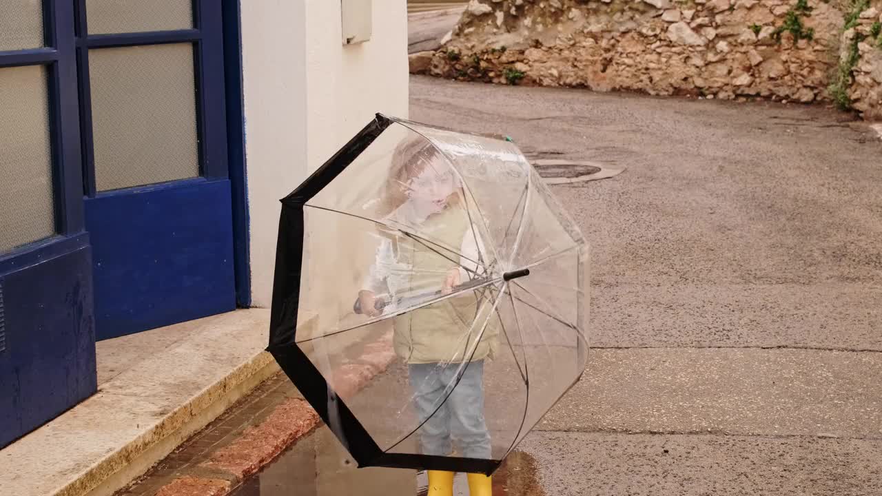 可爱的金发小女孩穿着黄色胶靴，在秋天的雨天里在城市街道上玩着大伞。视频素材