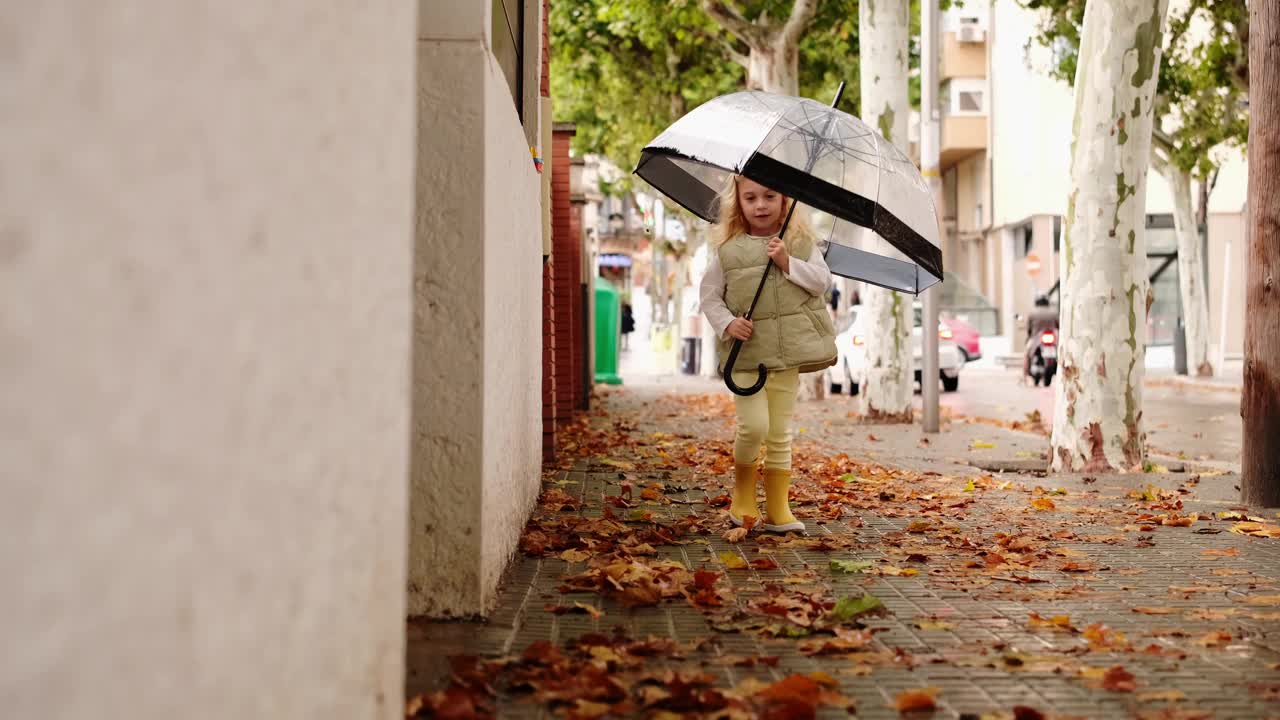 可爱的金发小女孩带着透明的大雨伞在雨天走在城市街道上。视频素材