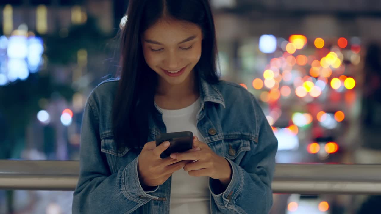 美丽的年轻亚洲女子站在城市街道在晚上使用手机。一个快乐的女人站在那里用手机打字视频素材