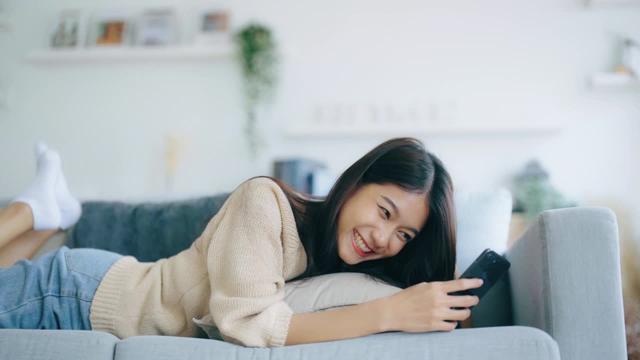 快乐的年轻亚洲女人在家里舒适的沙发上放松地用智能手机发短信，微笑的女孩用手机，在小工具上浏览无线互联网，在家网上购物视频素材