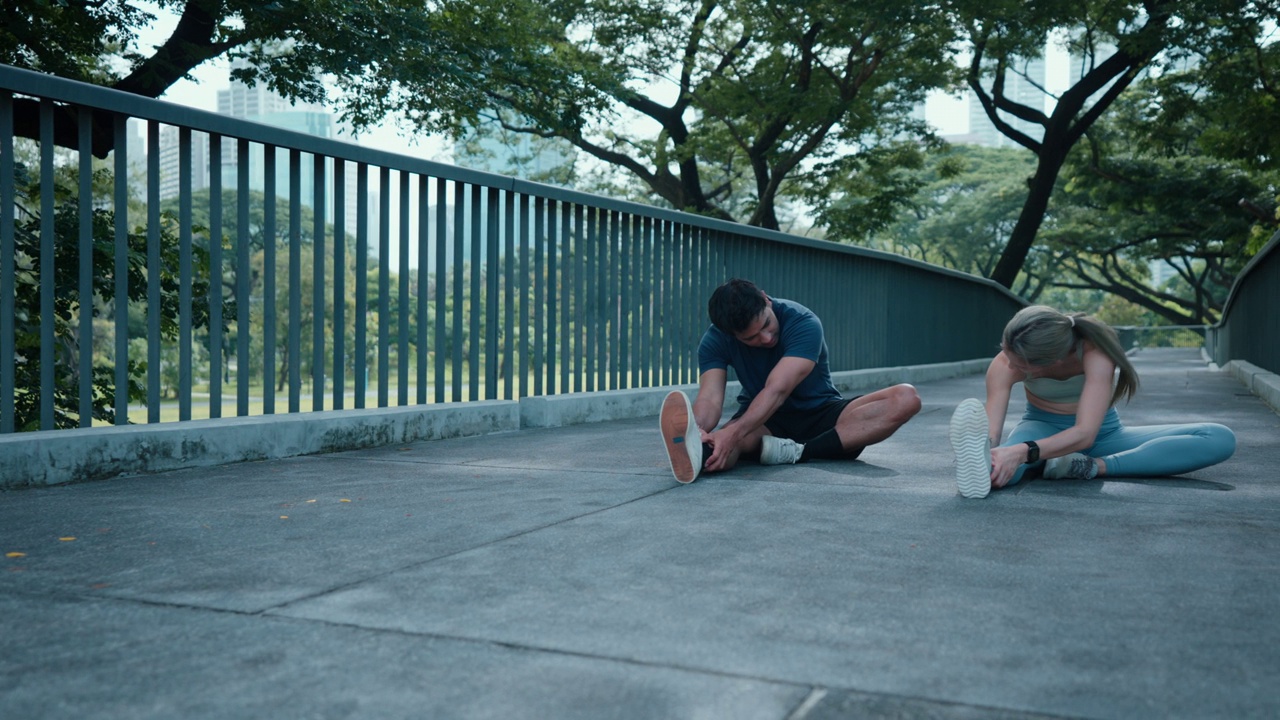 图为，一对穿着运动服的亚洲青年夫妇在公园慢跑结束后，为了减少受伤，正坐在地板上伸展腿部。视频素材