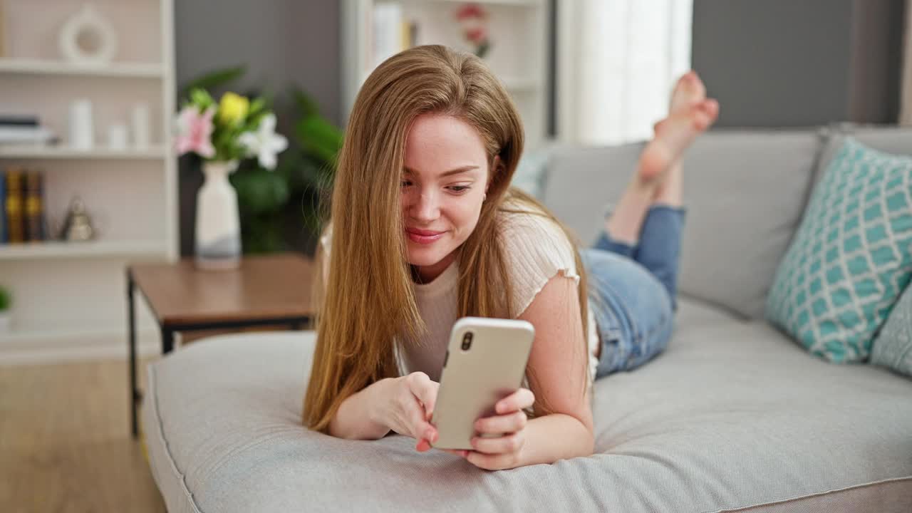 年轻的金发女郎躺在家里的沙发上用智能手机自拍视频素材