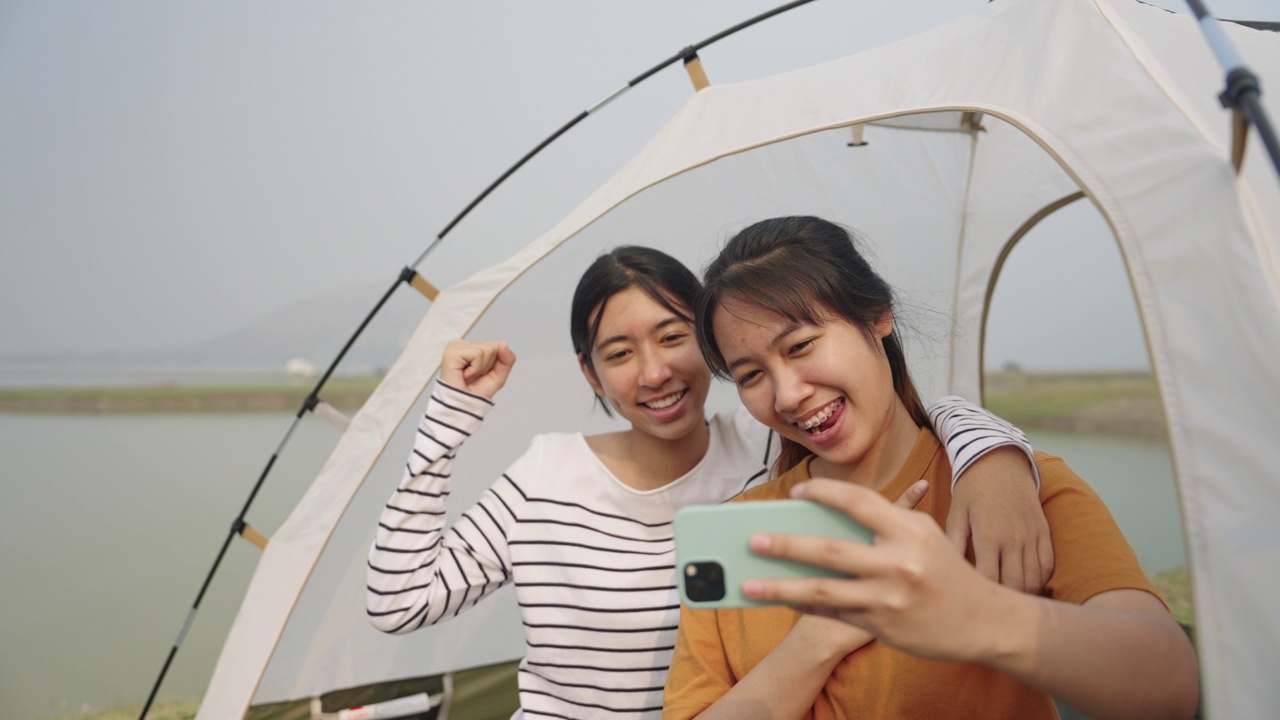 两个亚洲女孩坐在帐篷里用智能手机自拍，一起在户外露营视频素材