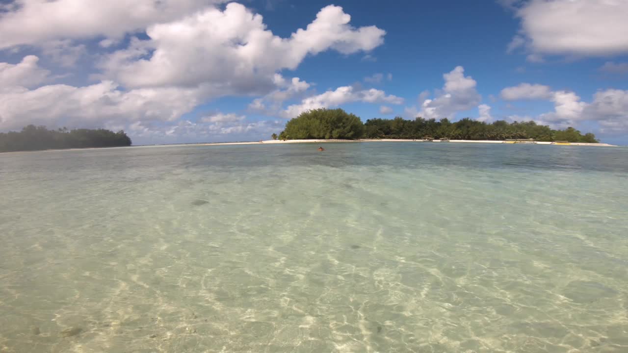 从Muri海滩向外看Koromiri岛的仍然视图站在水中，蛤蜊放松的海盐海洋绿松石泻湖轻轻地拍打着海边，蓝天和云层反射在水面上视频素材