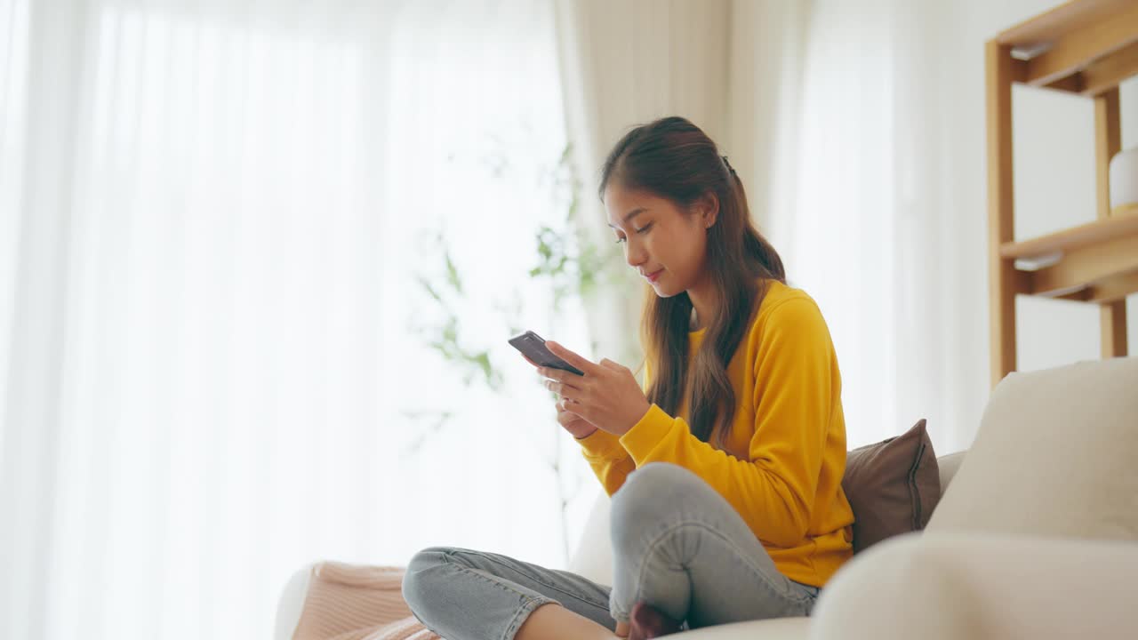 快乐的年轻亚洲女人在家里舒适的沙发上放松，在智能手机上输入聊天信息，微笑的女孩用手机聊天，在浏览器上搜索信息，无线互联网，网上购物视频素材