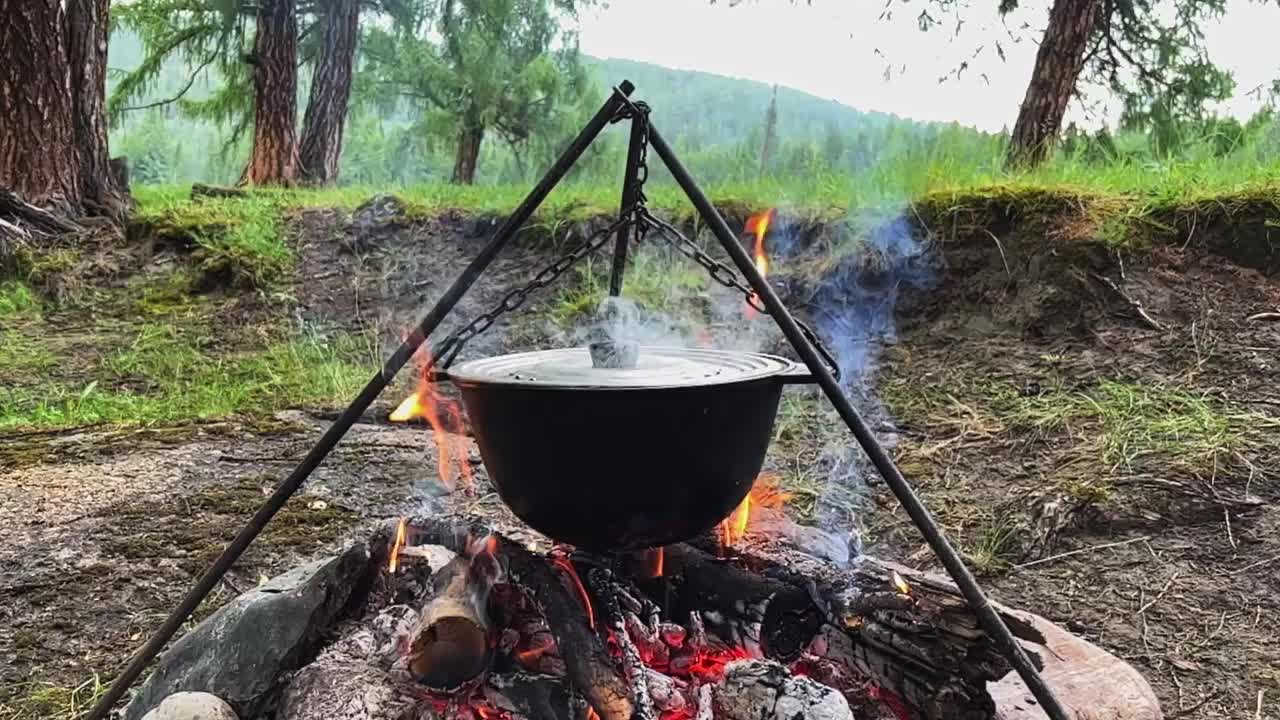 游客们在篝火上的露营锅里准备午餐。在户外山河岸边的森林里做饭。旅行营地，营火地点。视频下载