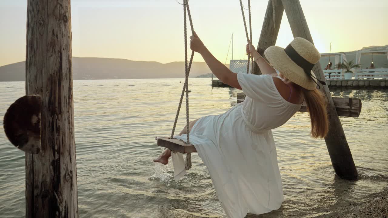 慢动作的海滩绳秋千，一个女人在一个飘逸的裙子品味摇摆在日落。旅游的精神，夏日的诱惑，对自由的拥抱，以及对旅行的探索。视频素材