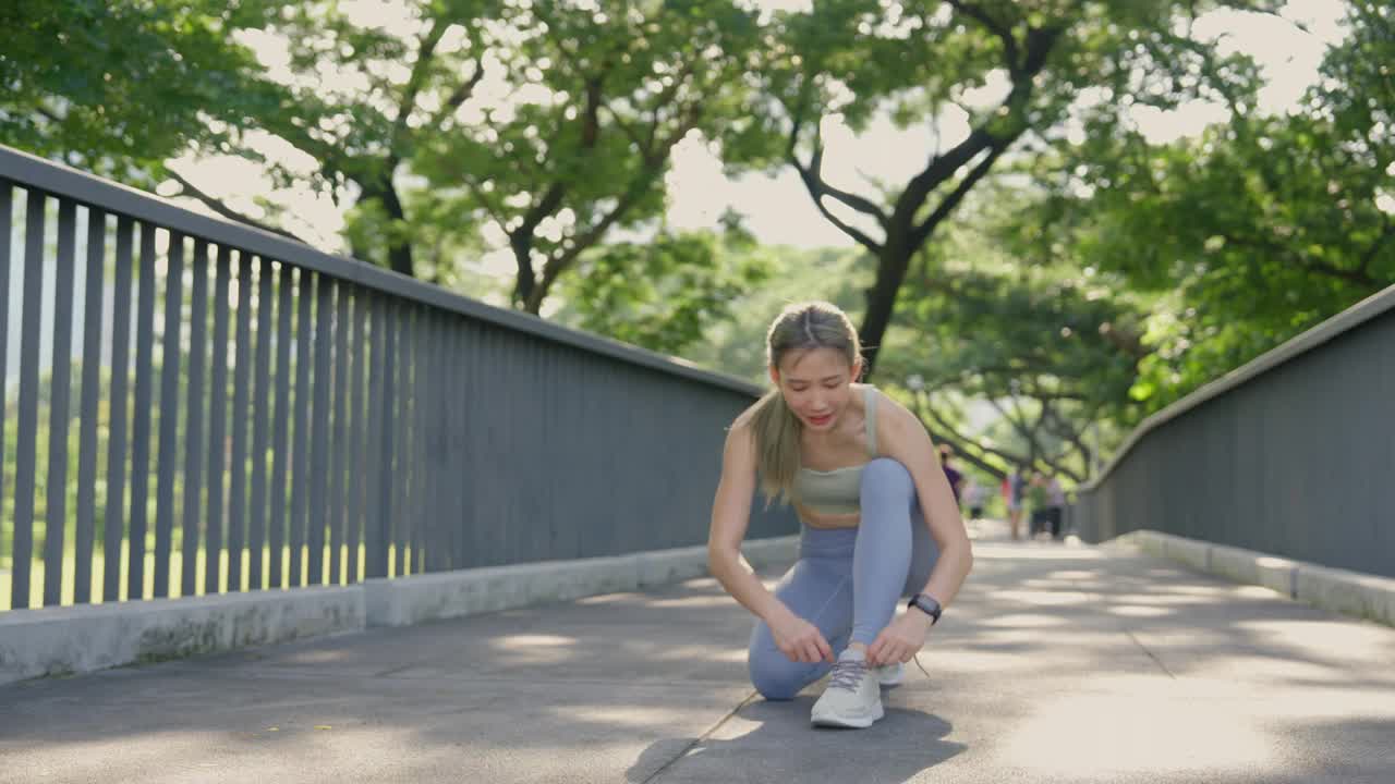 在一个阳光明媚的公园里，一名年轻的亚洲女子系好鞋带，准备在树木环绕的高架人行道上慢跑。视频素材