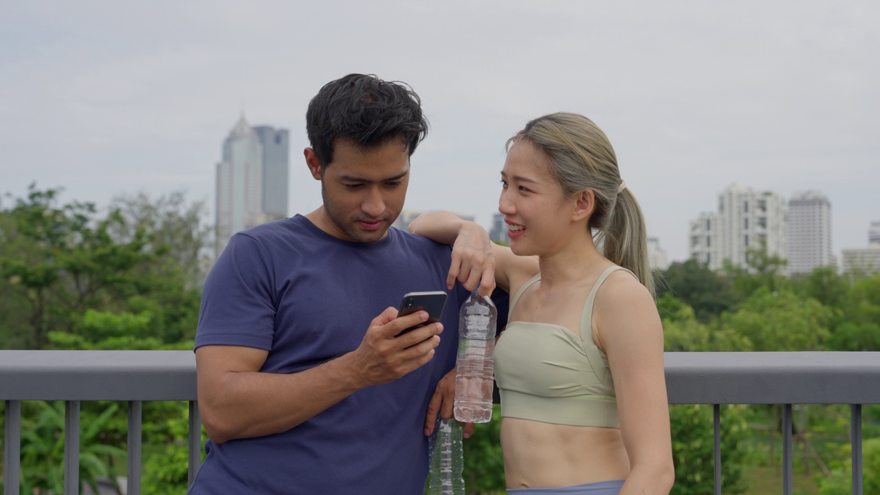 一对穿着运动服的年轻亚洲夫妇一起在智能手机上翻看照片，他们在市中心的一个公园里一起慢跑后微笑着。视频素材