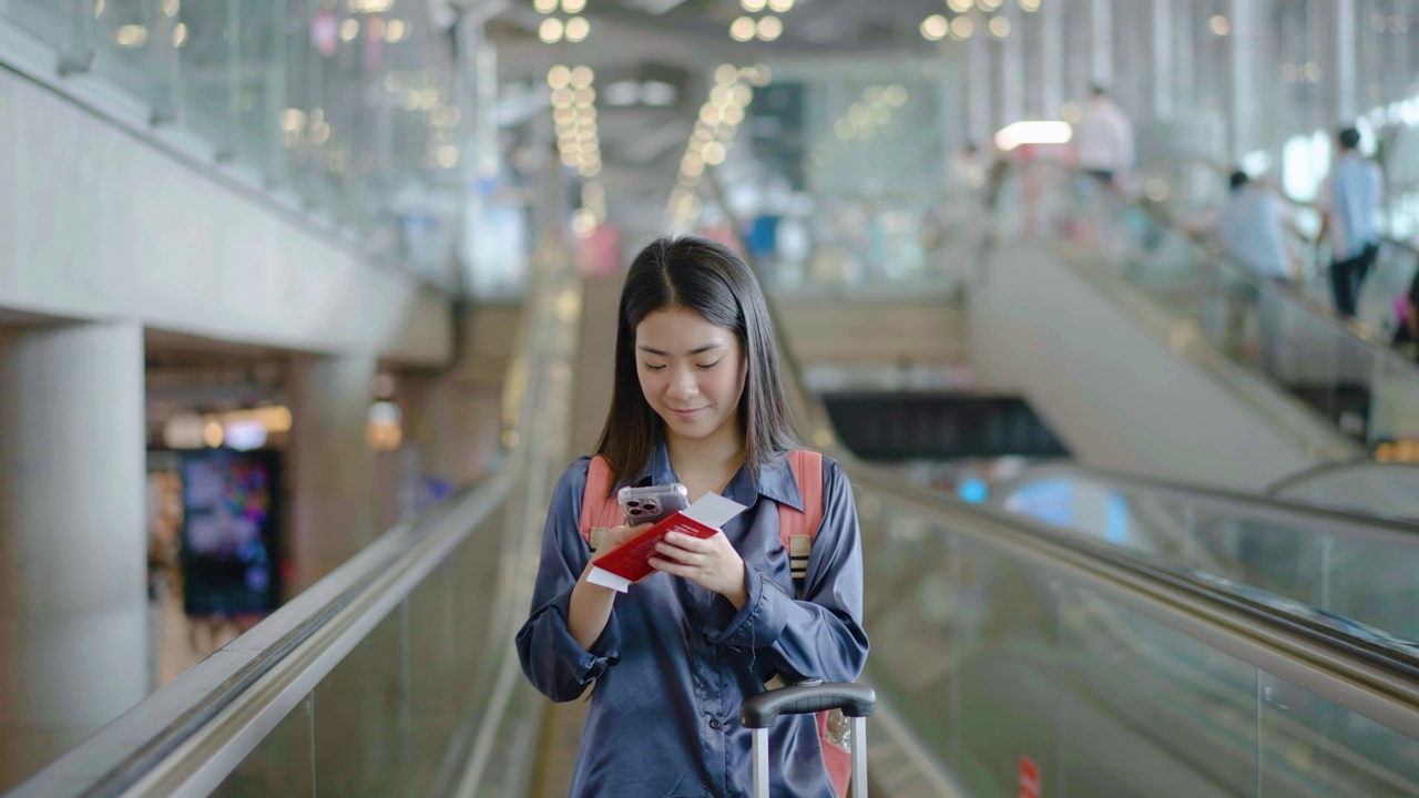 年轻的亚洲女子在机场候机楼的自动扶梯上检查手机。女性在旅途中提着行李。女孩用智能手机聊天，微笑着等飞机。旅行的概念视频素材