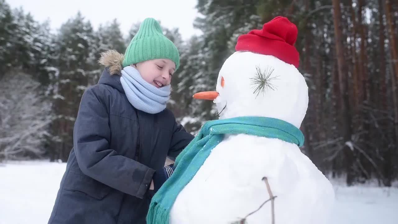 孩子和雪人说话，给他讲故事。男孩堆了一个雪人，一个快乐的孩子在白雪皑皑的冬日森林里玩耍，为雪人拉直了围巾和帽子视频素材