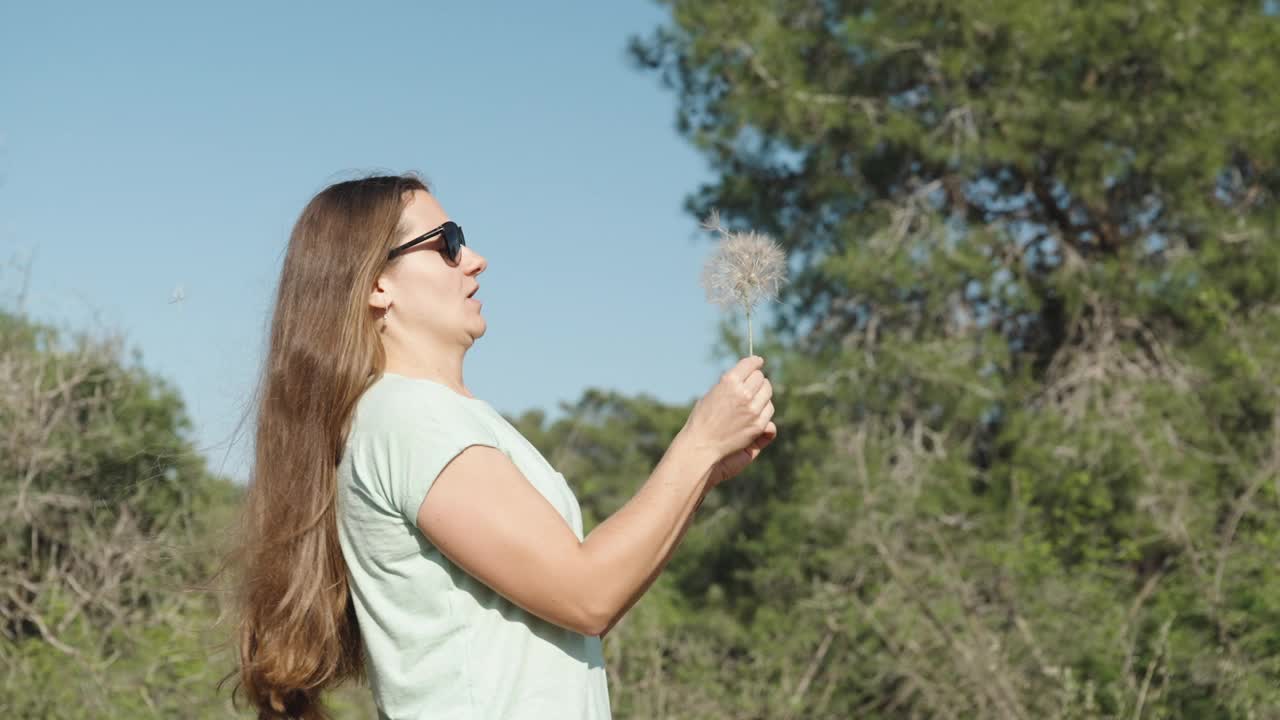 一名妇女吹着一颗大而圆的角鹫花成熟的种子，在蓝天和树木的映衬下，它们在风中向她的脸上旋转。缓慢的运动。视频素材