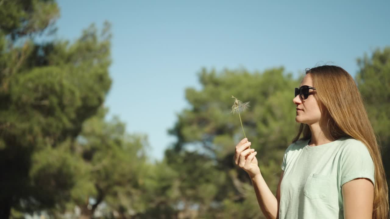 慢镜头中，女人对着金角花吹气，在蓝天和树木的映衬下，所有的种子随风散落。视频素材