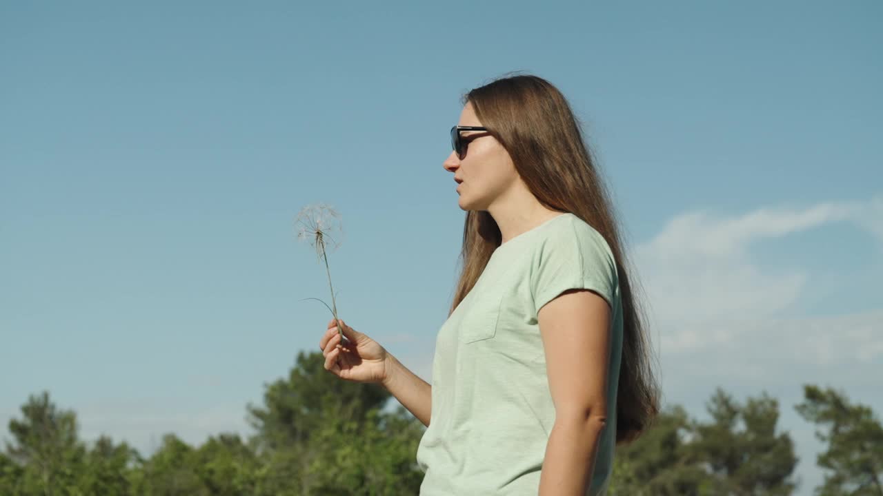 一位长发女子在空中吹着一颗角鹫花的种子，它们在蓝天的背景下随风散落。缓慢的运动。视频素材