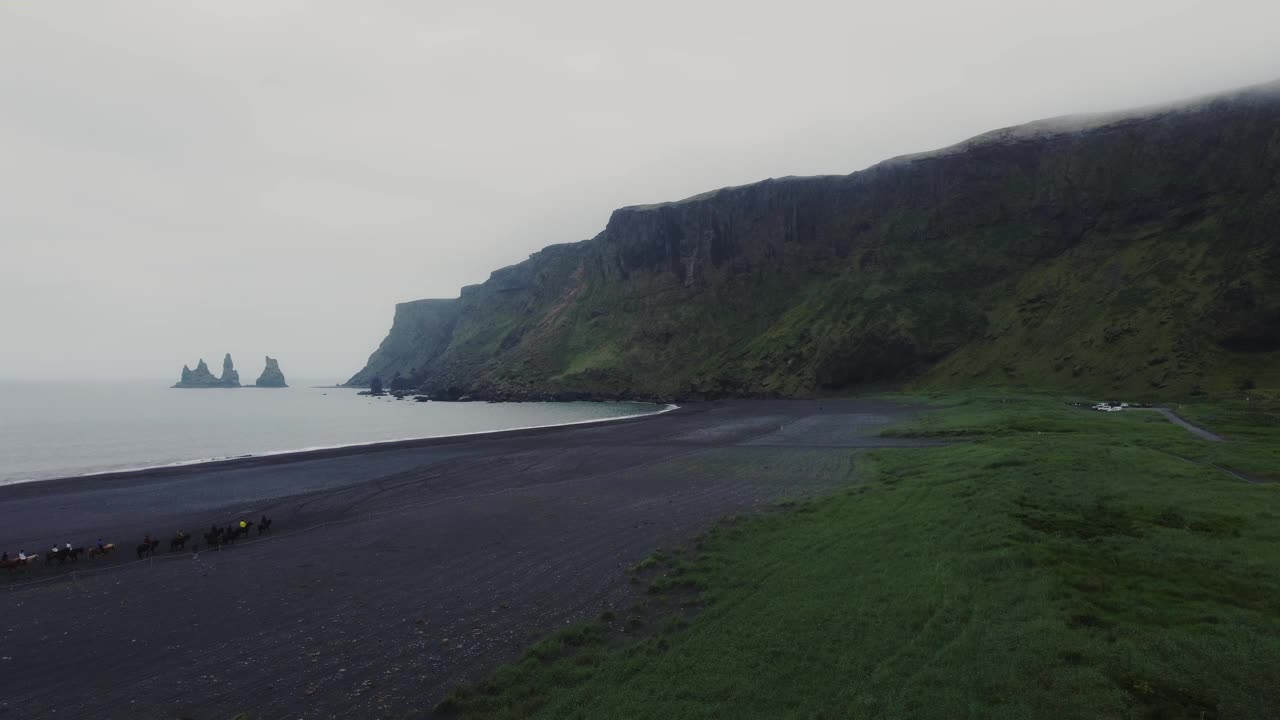 冉冉升起的无人机拍摄到美丽的蓝色海浪冲击着冰岛的黑色沙滩，远处有一群冰岛马和巨大的岩层视频素材