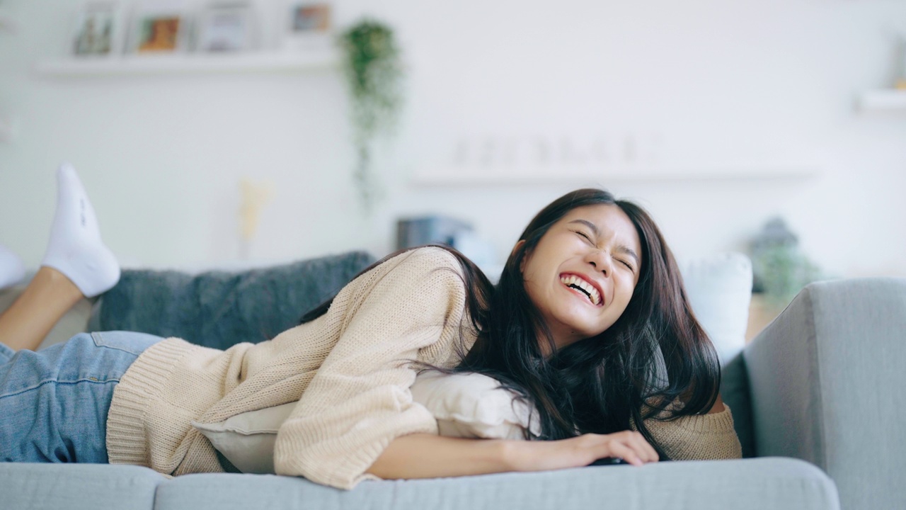 快乐的年轻亚洲女人在家里舒适的沙发上放松地用智能手机发短信，微笑的女孩用手机，在小工具上浏览无线互联网，在家网上购物视频素材