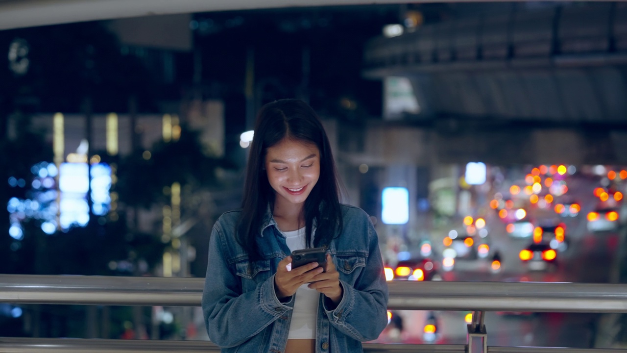 美丽的年轻亚洲女子站在城市街道在晚上使用手机。一个快乐的女人站在那里用手机打字视频素材