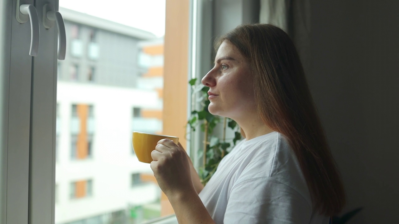 美丽放松的女人，一杯茶或咖啡，坐在家里的窗台上看着窗外视频素材