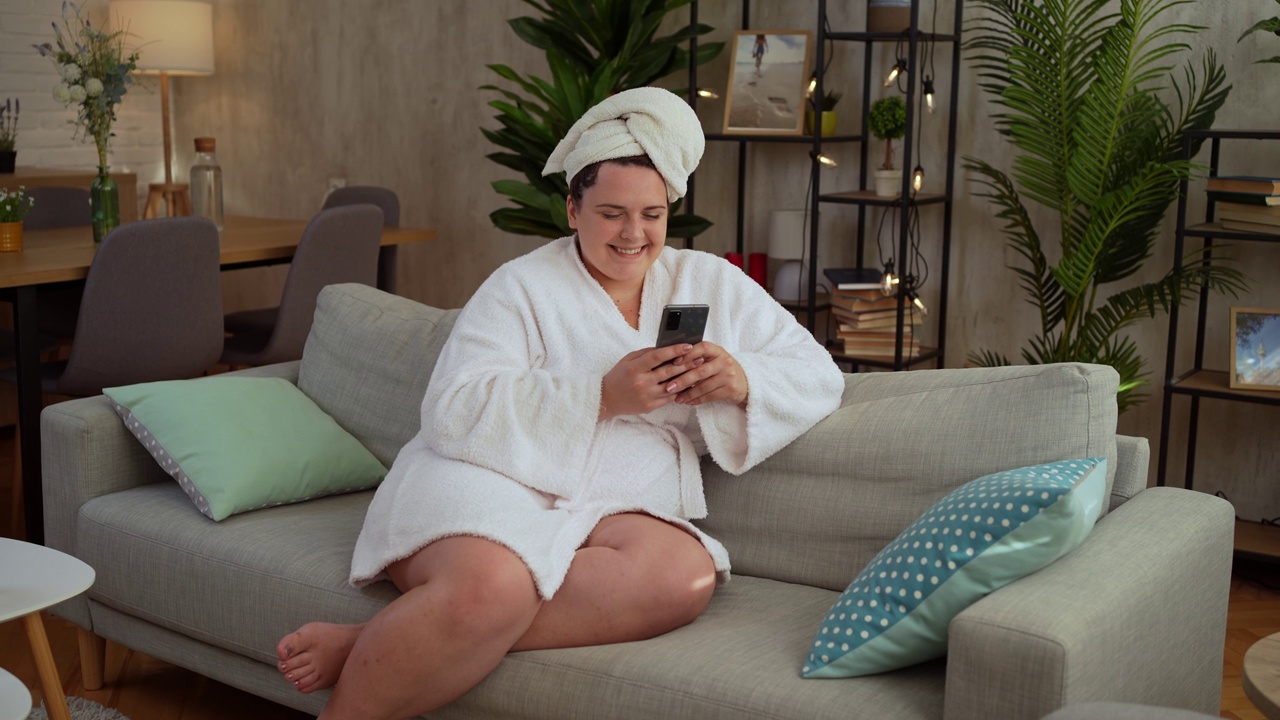 穿着浴袍的年轻女子在客厅玩手机视频素材