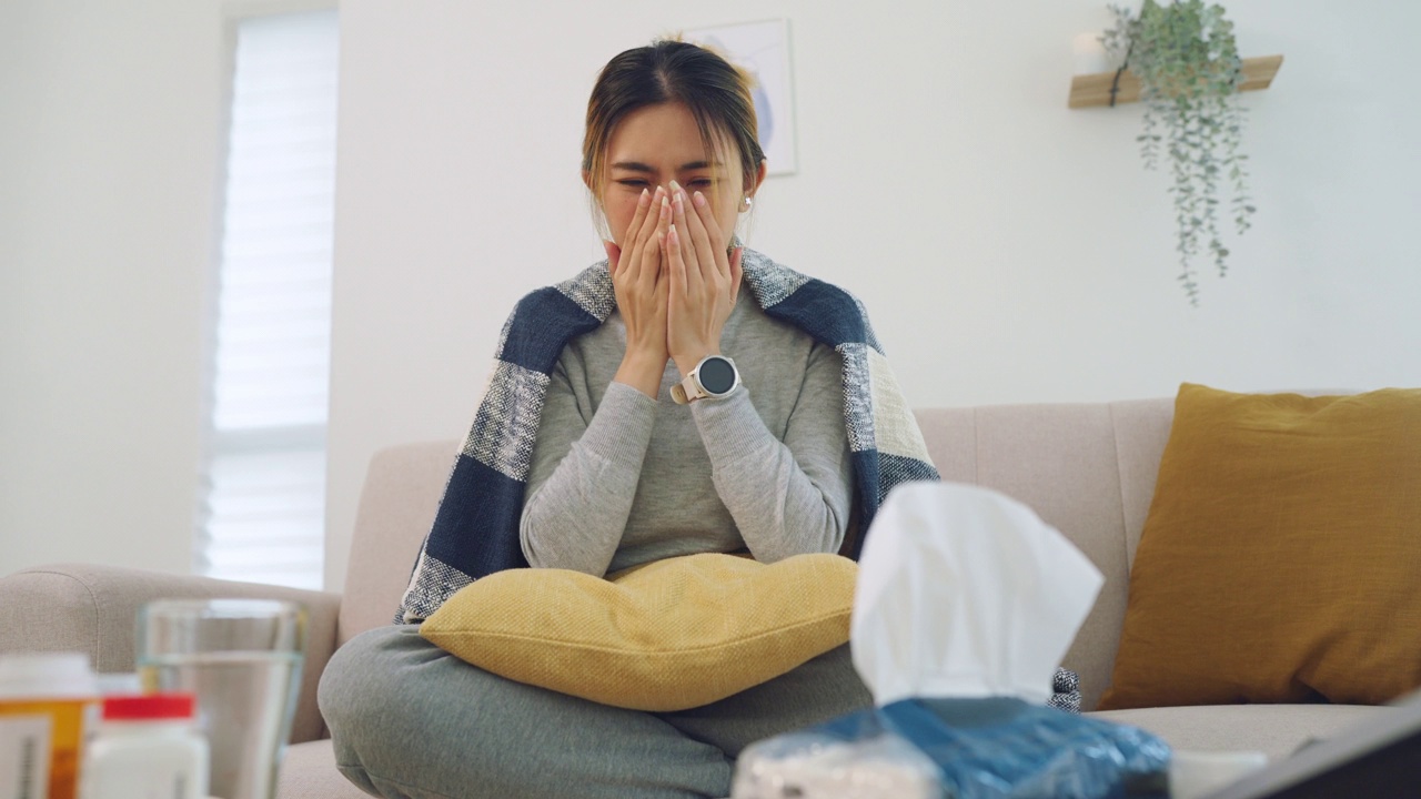 亚洲妇女有流感病毒覆盖格纹在纸巾打喷嚏流鼻涕坐在家里的沙发上恶心，季节性病毒视频素材